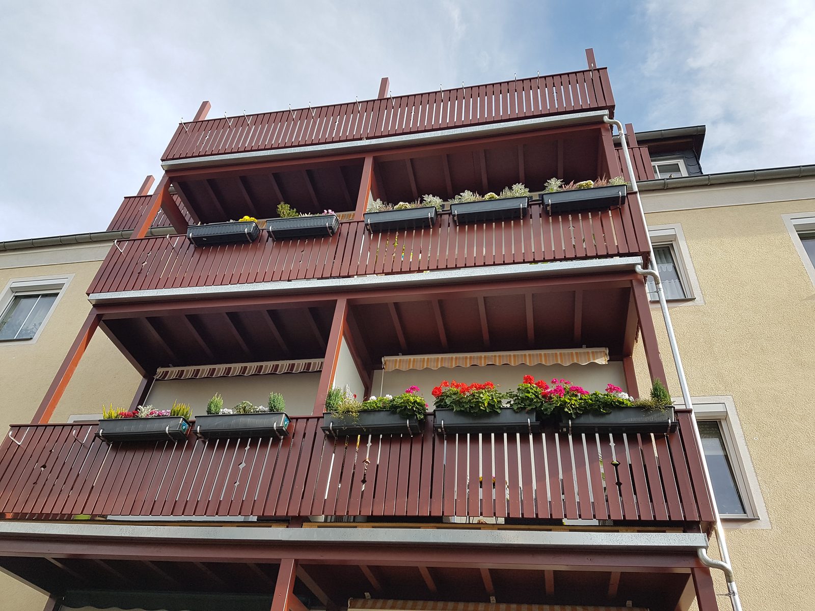 Balkonanlage aus rotbraunem Holz über drei Geschosse, mit bepflanzten Blumenkästen